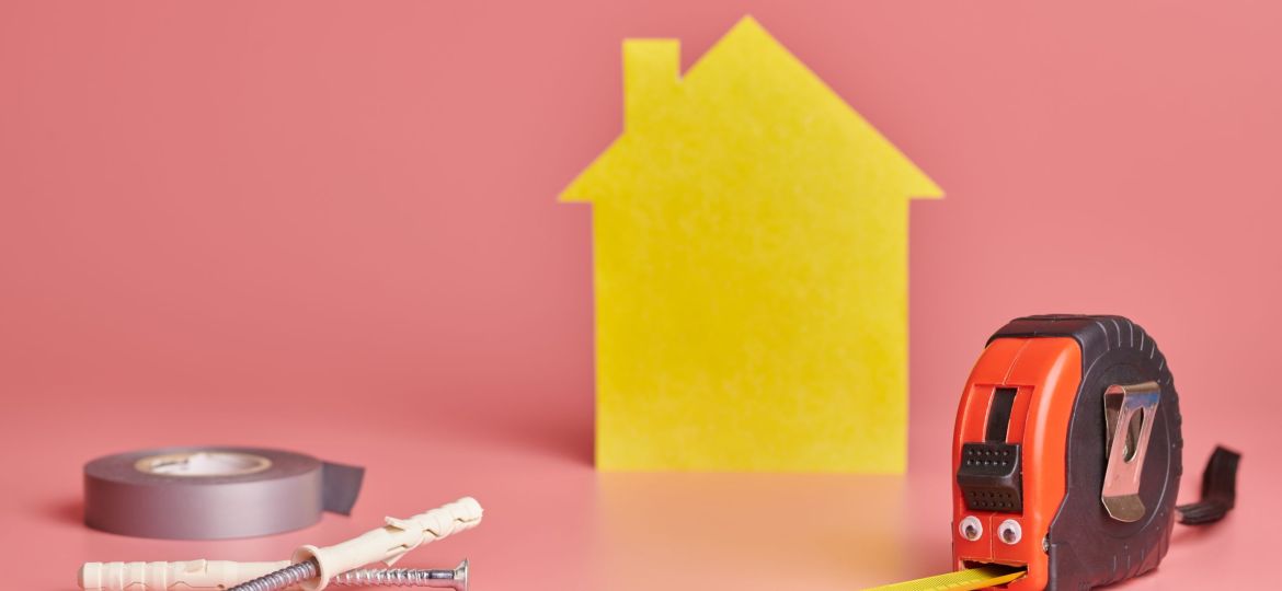 House renovation funny concept. Metal tape measure and other repair items. Home repair and redecorated concept. Yellow house shaped figure on pink background. (Demo) (Demo)