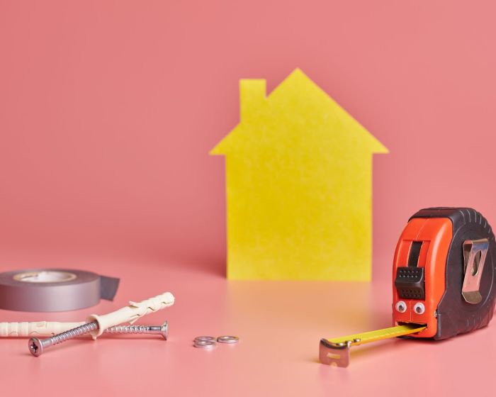 House renovation funny concept. Metal tape measure and other repair items. Home repair and redecorated concept. Yellow house shaped figure on pink background. (Demo) (Demo)